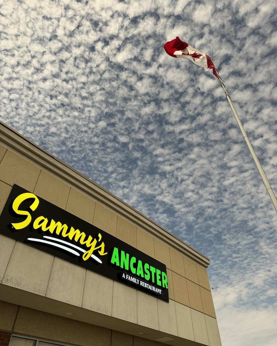 The lit Sammy's Ancaster sign on the storefront, Canadian flag flying against a bright cloud-flecked sky